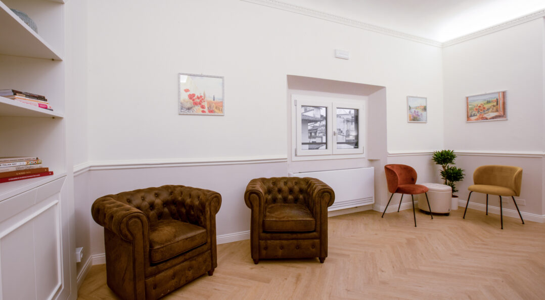 Common lounge area at Hotel Settefiori featuring two brown chesterfield armchairs, modern colorful chairs, light wood flooring, and floral paintings on the walls.